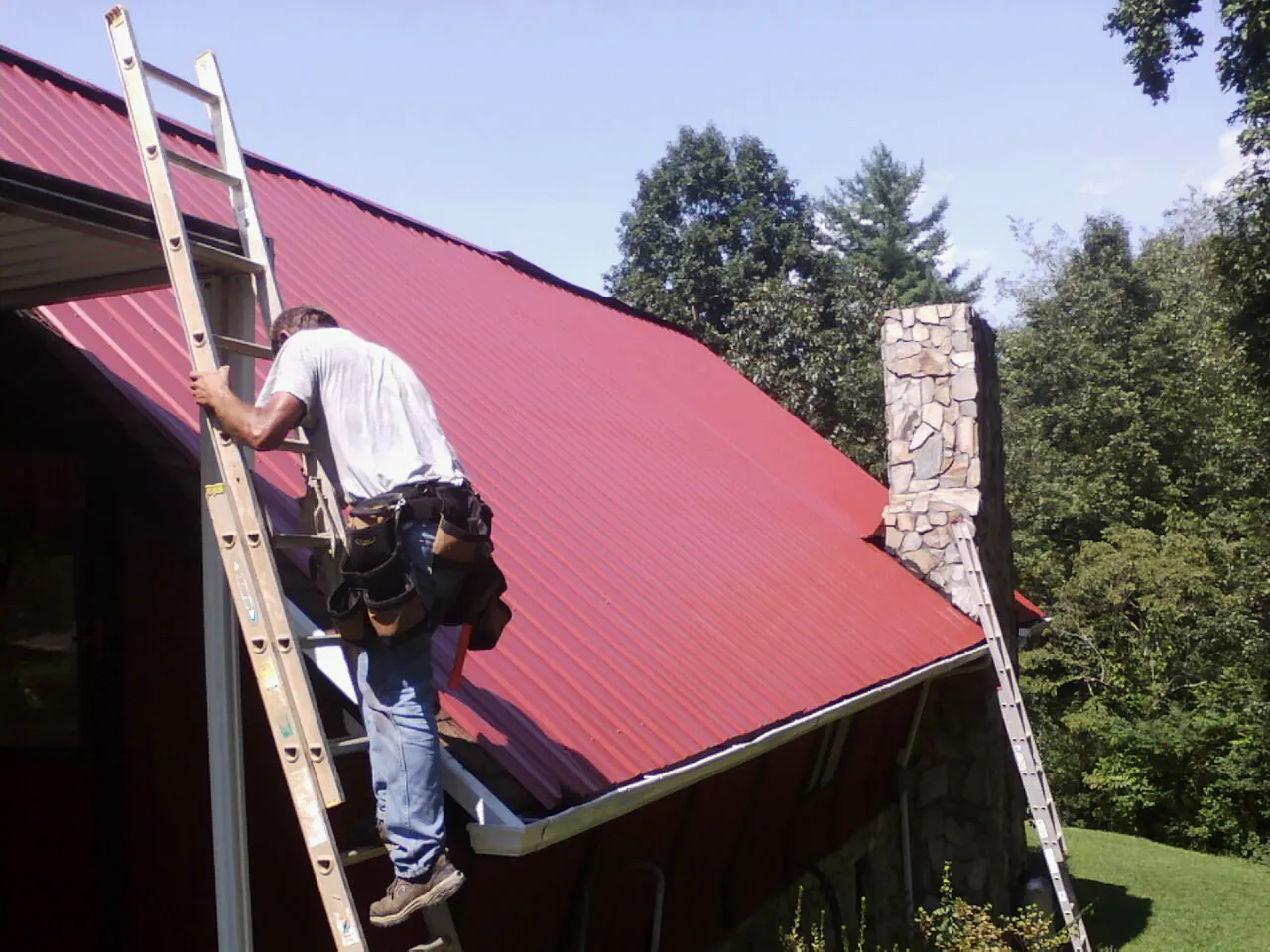 Roofer on a ladder beside a red metal roof and stone chimney — Soffit & Fascia Repair by Roofing Brothers in Solon